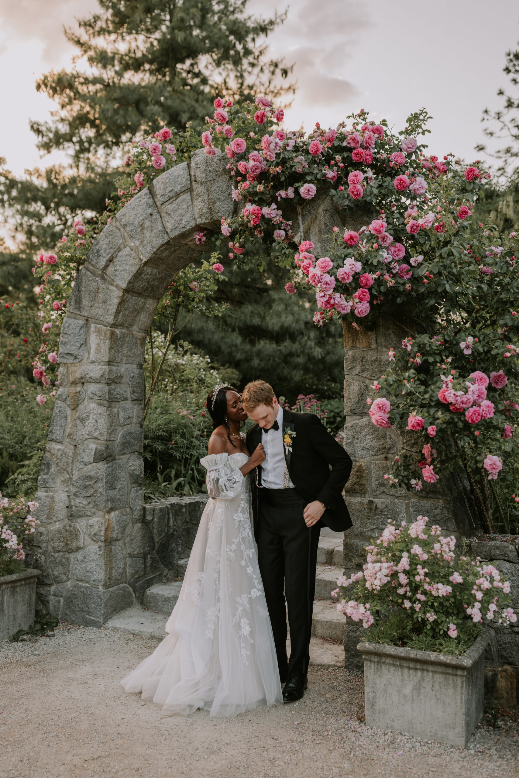 elegant Couple standing in front of stone archway covered in roses pink and colourful roses